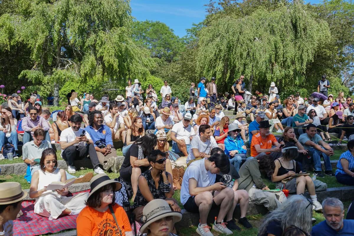 A diverse group of people sitting on grassy steps outdoors, some reading or using phones, under sunny skies and surrounded by leafy trees.
