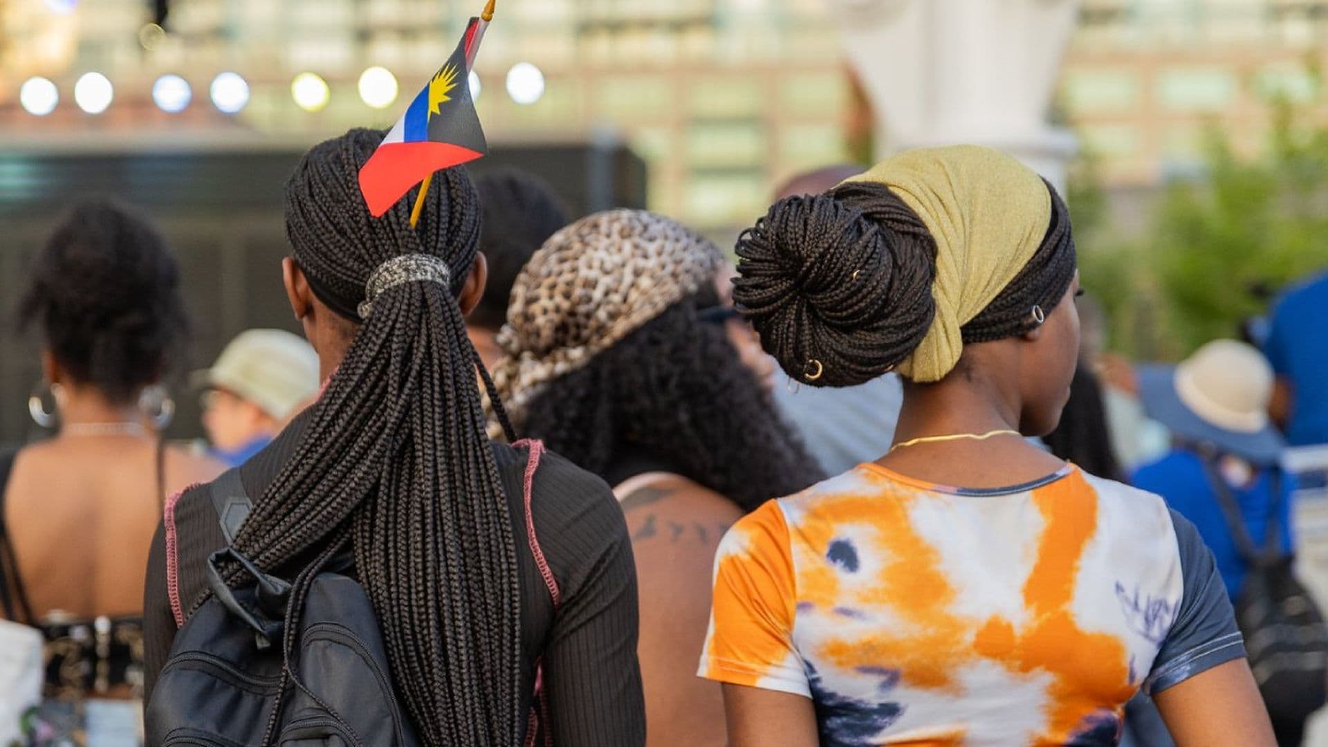 People at an outdoor event, one with a braided hairstyle and flag, another with a headwrap, against a blurred backdrop of lights.