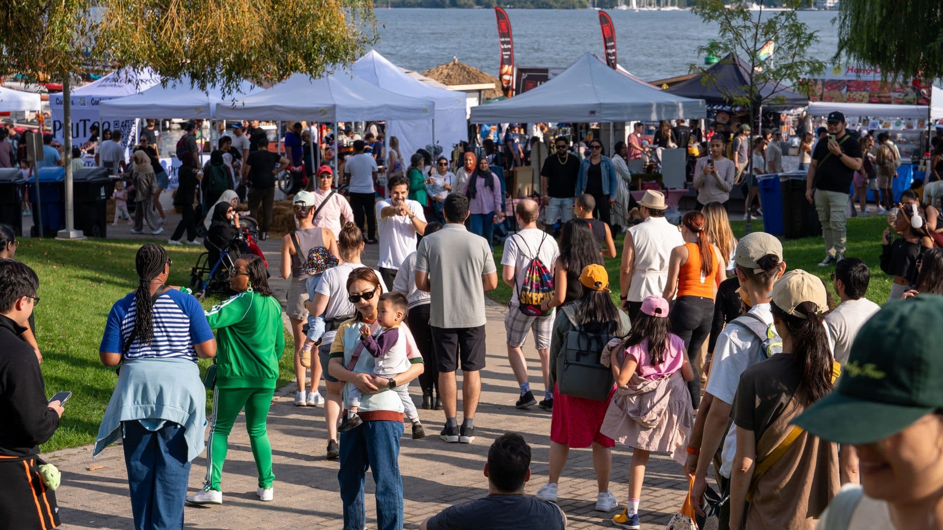 A lively outdoor festival by a waterfront, with diverse crowds exploring vendor booths under white tents on a sunny day.